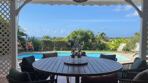 Ein überdachter Außenbereich mit Blick auf einen Pool, ausgestattet mit einem runden Holztisch und Korbstühlen.
