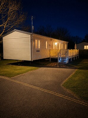 A single-story mobile home with a covered porch and a small deck, surrounded by a paved driveway and grassy areas. - Singh&Leite Family Caravan 10 (Clacton-on-Sea)