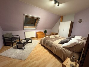 A bedroom with a bed, a chair, a table, a radiator, a window, and a wardrobe.