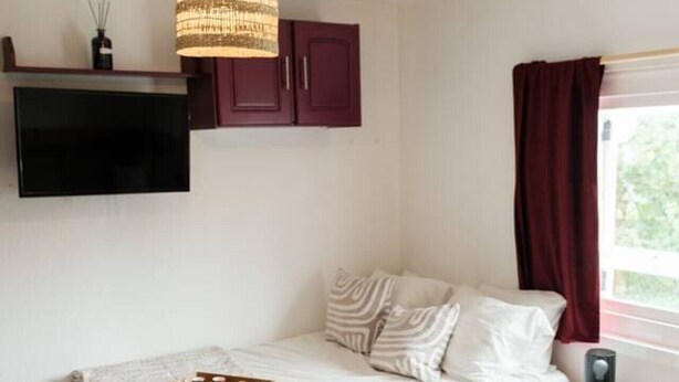 A bedroom with a bed, a bedside table with a lamp, a TV mounted on the wall, and a window with curtains. - Studio chaleureux avec vue montagne au cœur de Font-Romeu (Font-Romeu-Odeillo-Via)