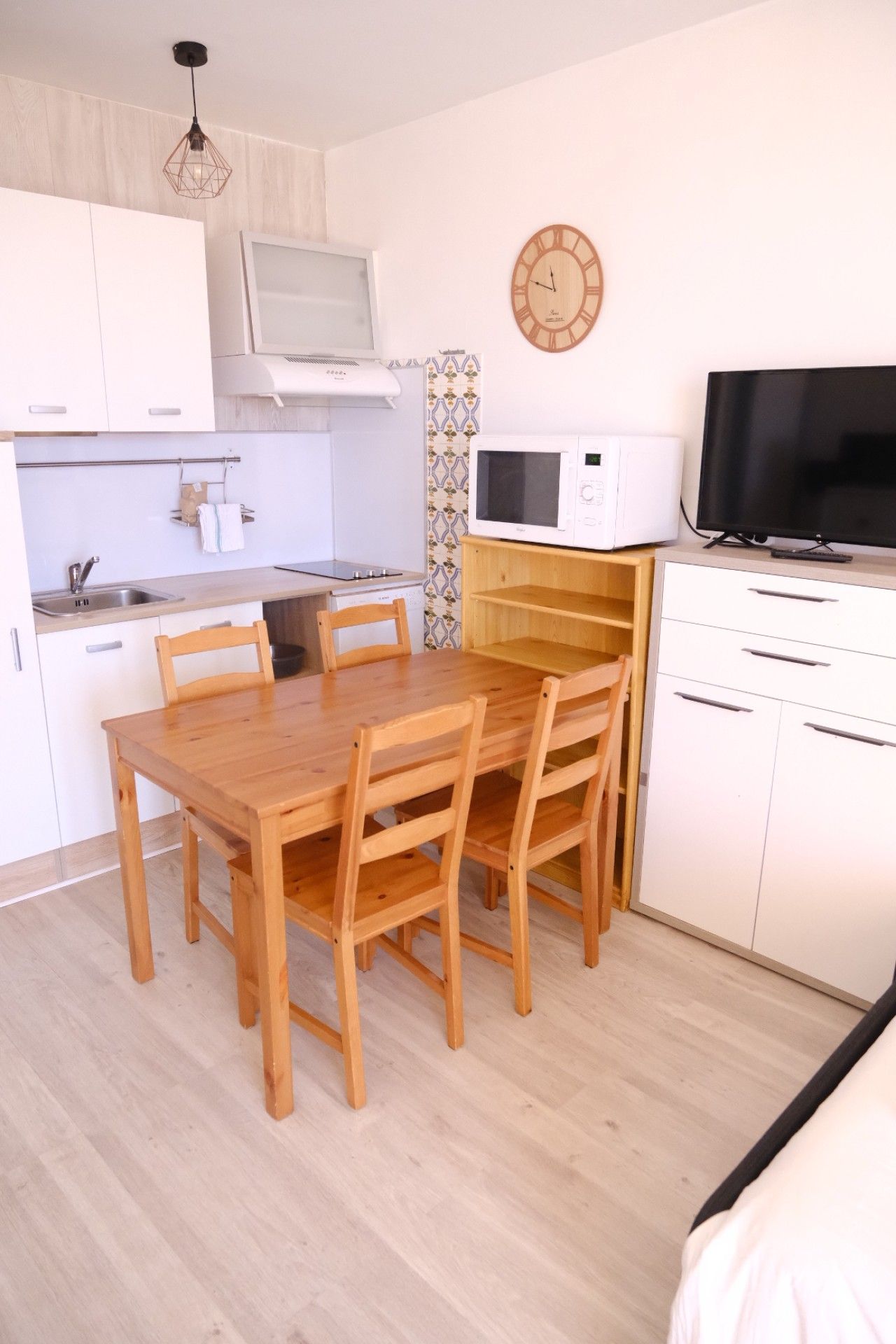 Een kleine keuken met een eettafel, stoelen, een magnetron, een televisie en een klok aan de muur.