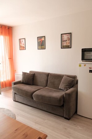 A living room with a brown sofa, a white refrigerator, and two framed pictures on the wall. - Studio cabine avec balcon ensoleillé pour 6 personnes en hyper-centre (Font-Romeu-Odeillo-Via)