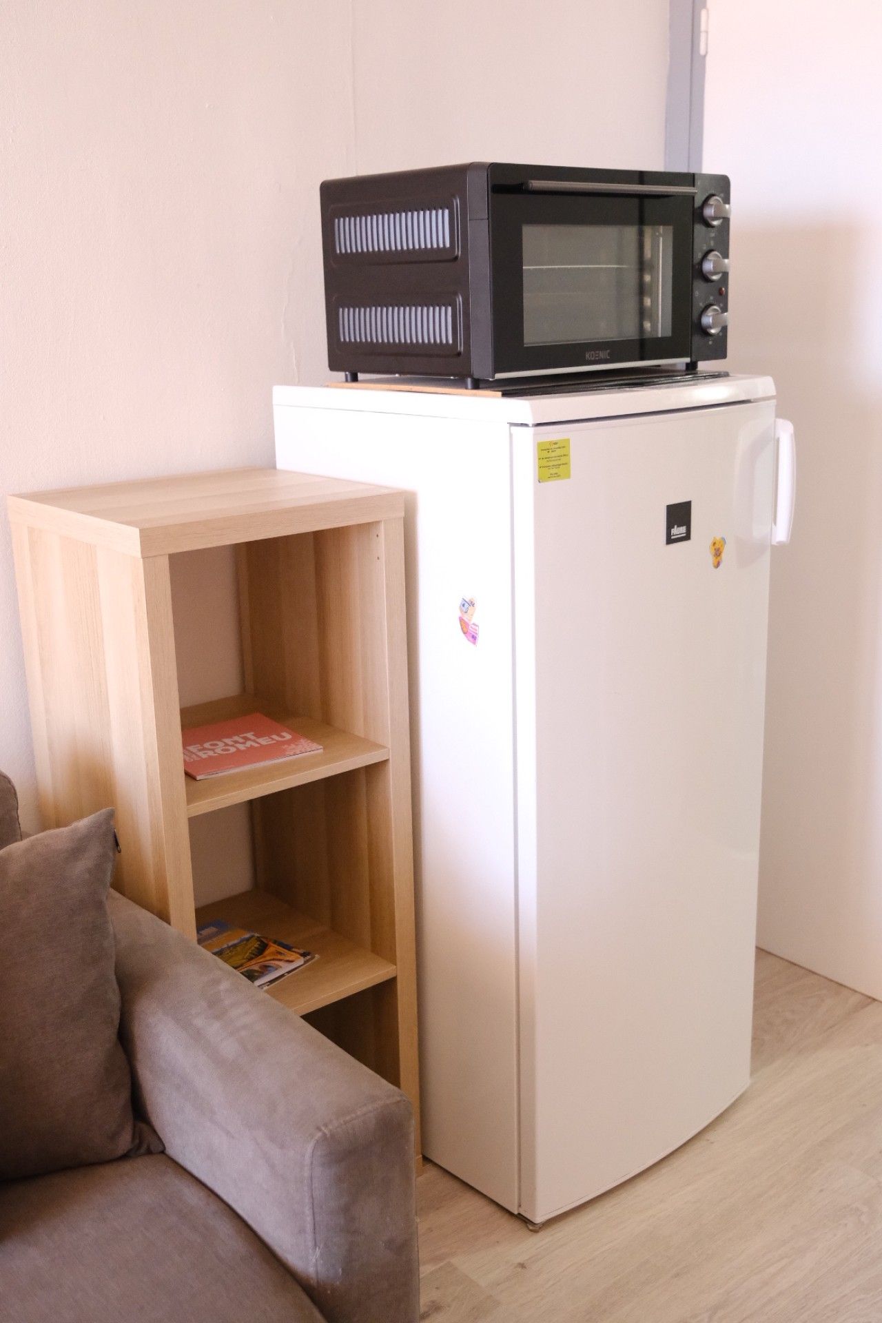 A compact living space with a white refrigerator, a microwave on top, a wooden shelf unit, and a brown sofa.