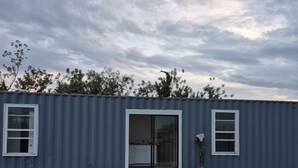 En envåningsbyggnad av metall med en träveranda, en skjutdörr i glas som ingång och ett litet utomhusområde med en skulptur av metall.
