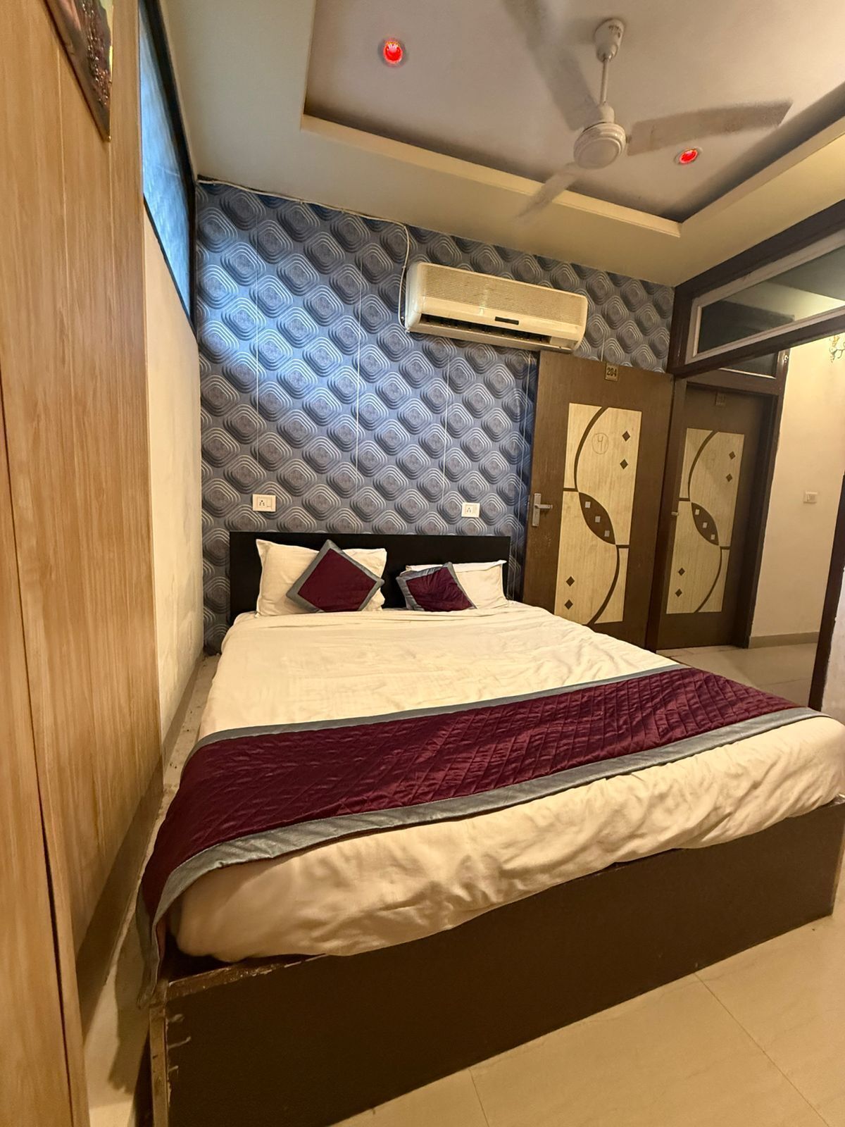 A hotel room with a bed, an air conditioning unit, a ceiling fan, and a patterned wall.