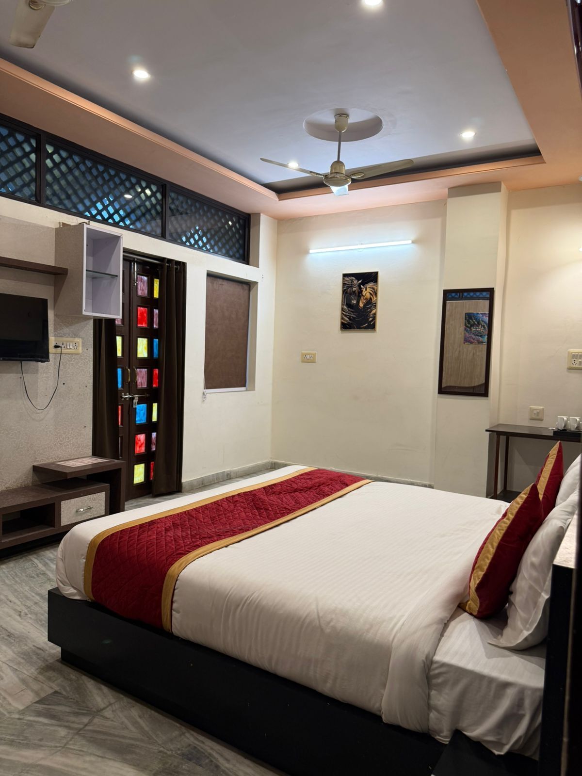 A hotel room with a bed, a ceiling fan, a TV, a desk, and artwork on the walls.