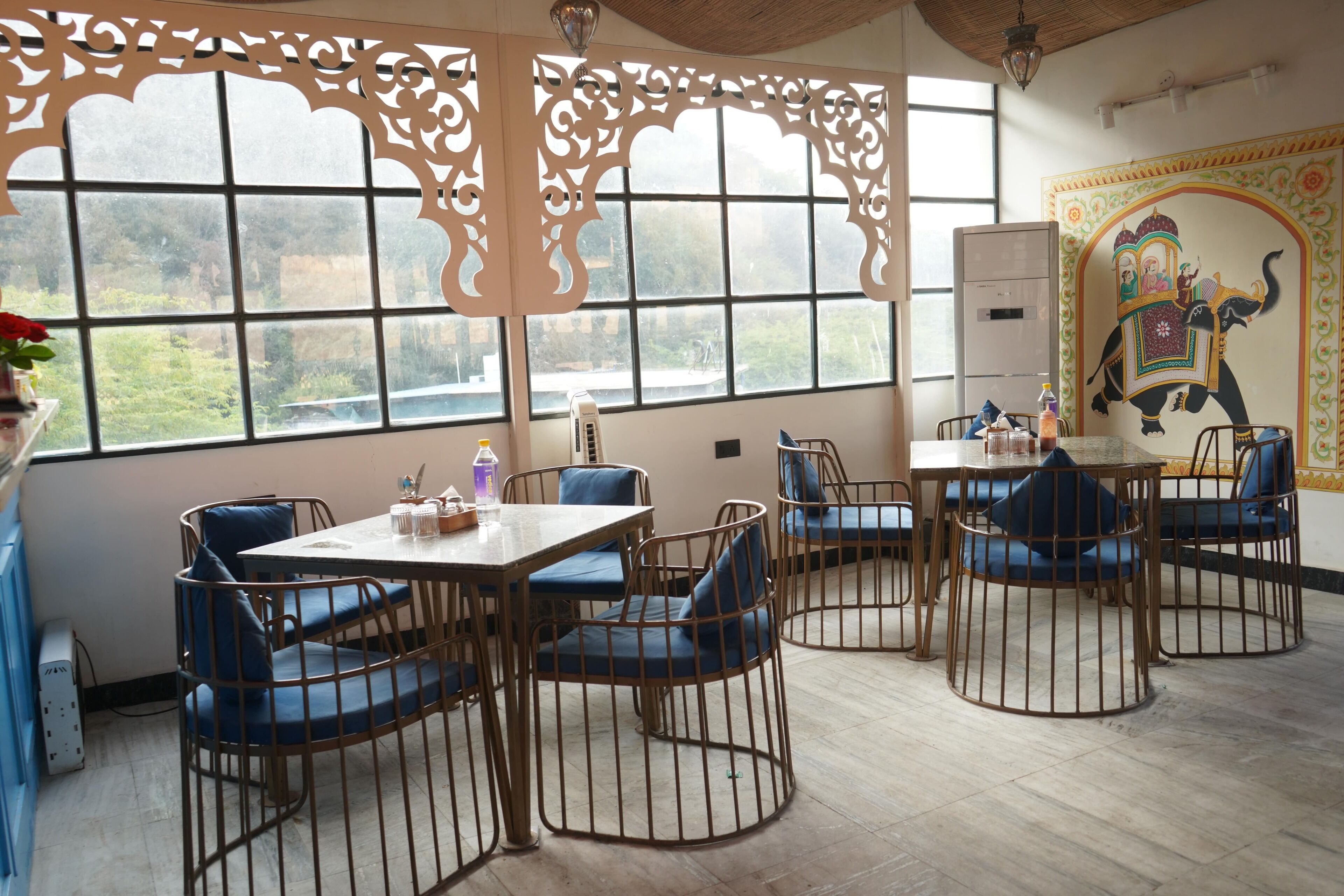 A dining area with round tables and blue cushioned chairs, large windows with decorative screens, and a mural of an elephant on the wall.