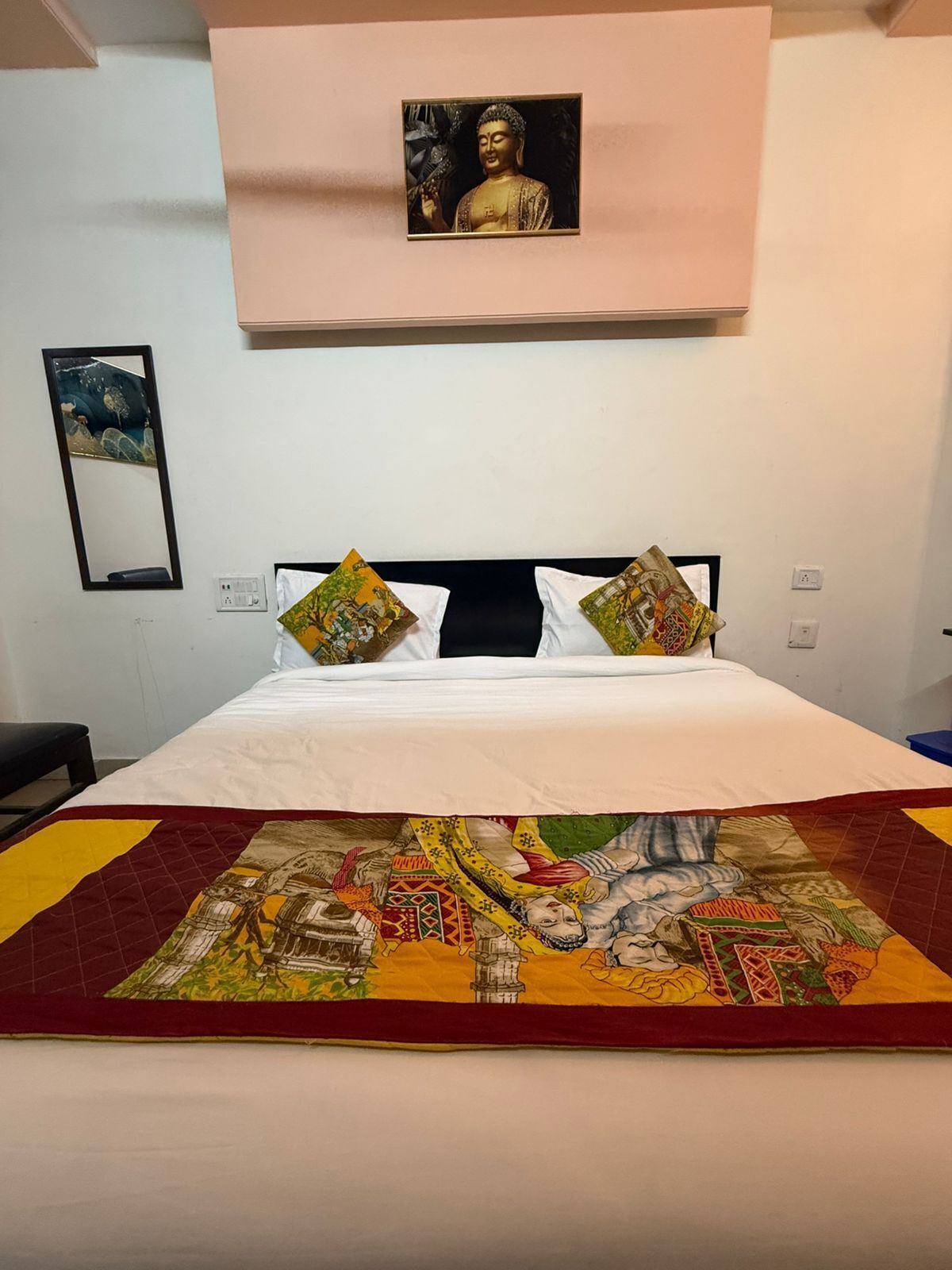 Een bed met witte beddengoed, twee kussens met kleurrijke patronen, een groot decoratief quilt, een ingelijste Boeddhabeeld aan de muur, en een spiegel met een schilderij.