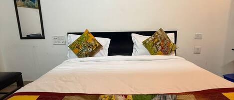 Een bed met witte beddengoed, twee kussens met kleurrijke patronen, een groot decoratief quilt, een ingelijste Boeddhabeeld aan de muur, en een spiegel met een schilderij.