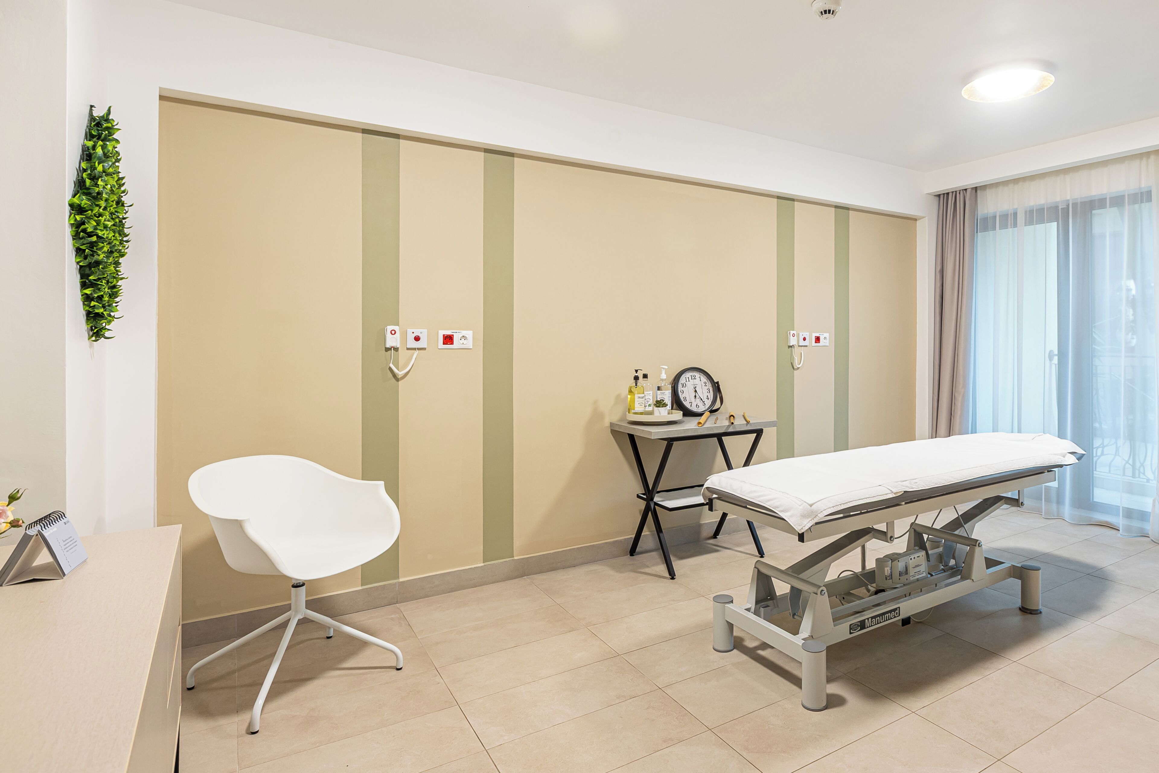 A clinical examination room with a white examination table, a white chair, a small table with a clock and medical supplies, and a green wall with vertical stripes.