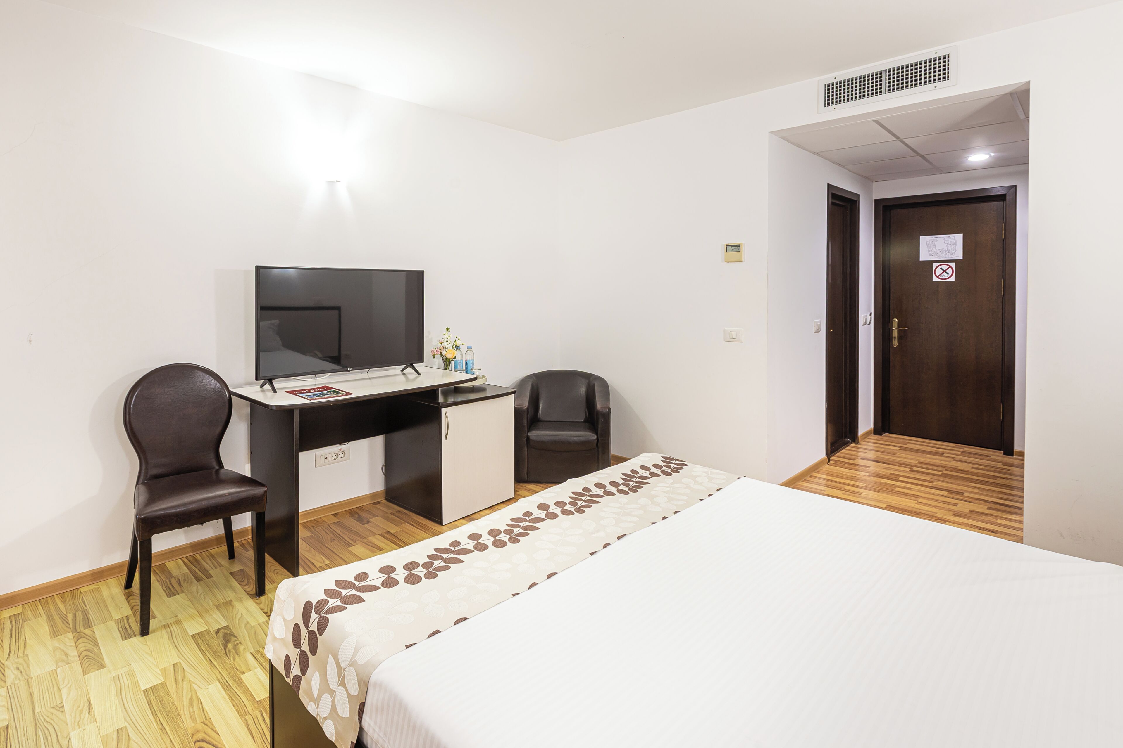 A hotel room with a bed, desk, two chairs, a television, and a door with a sign.