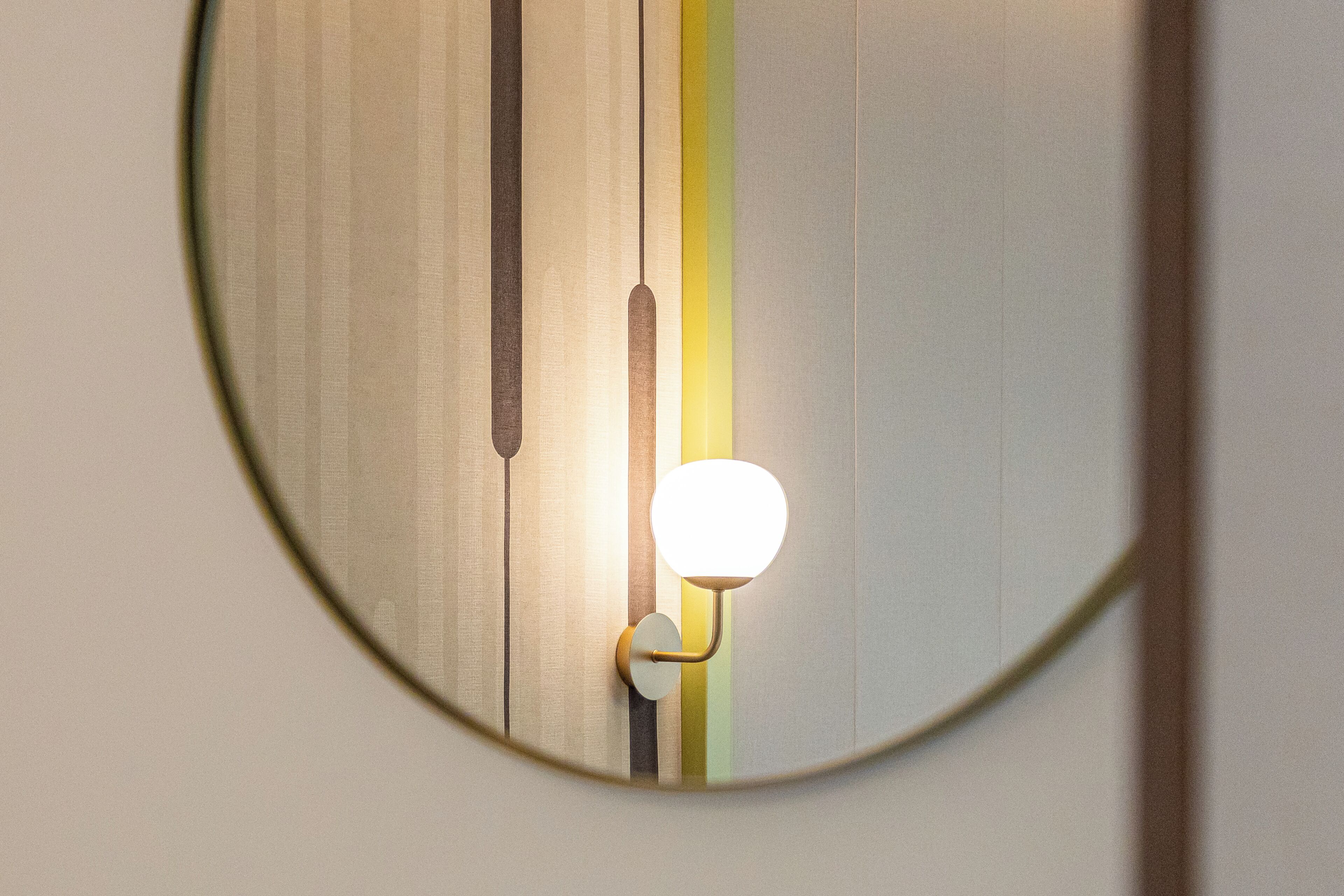 Un espejo circular que refleja una pared con rayas verticales y una lámpara.