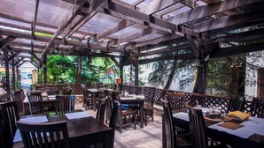An outdoor restaurant with wooden tables and chairs, covered by a wooden pergola, and surrounded by trees.