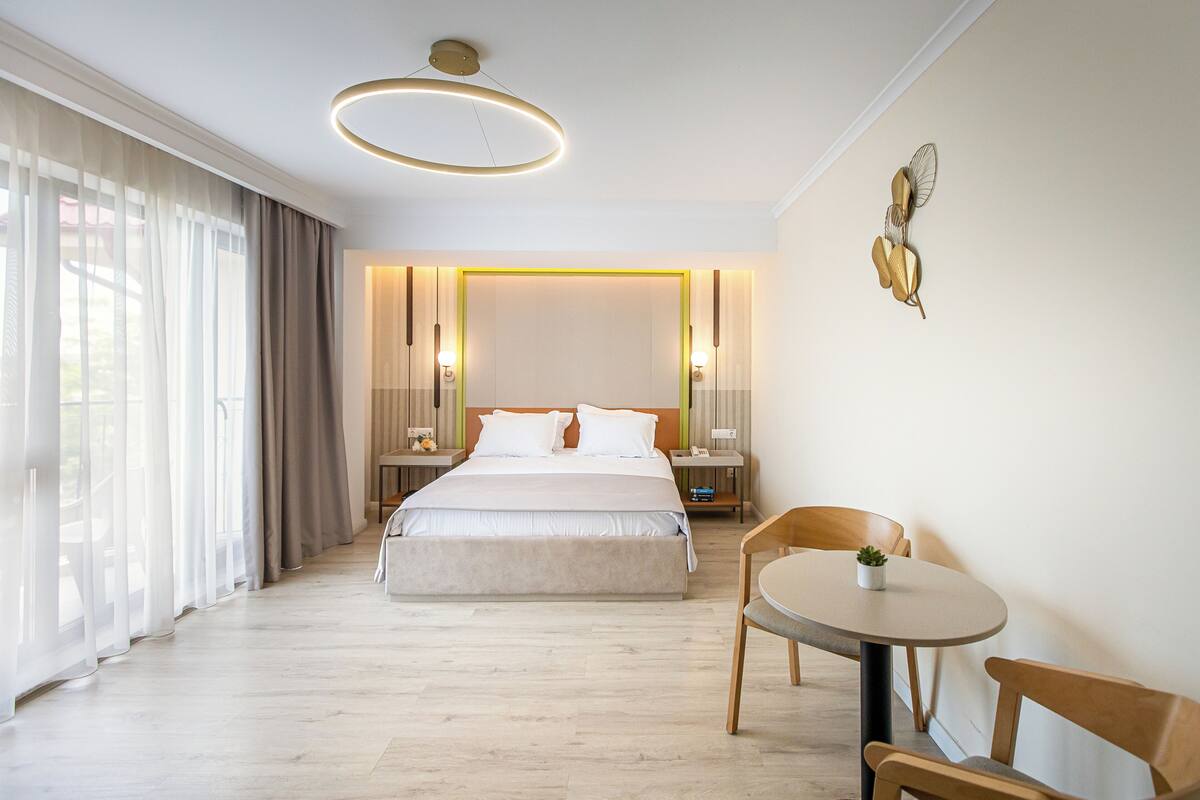 A hotel room with a large bed, bedside tables, a round table with chairs, and a view through sheer curtains.