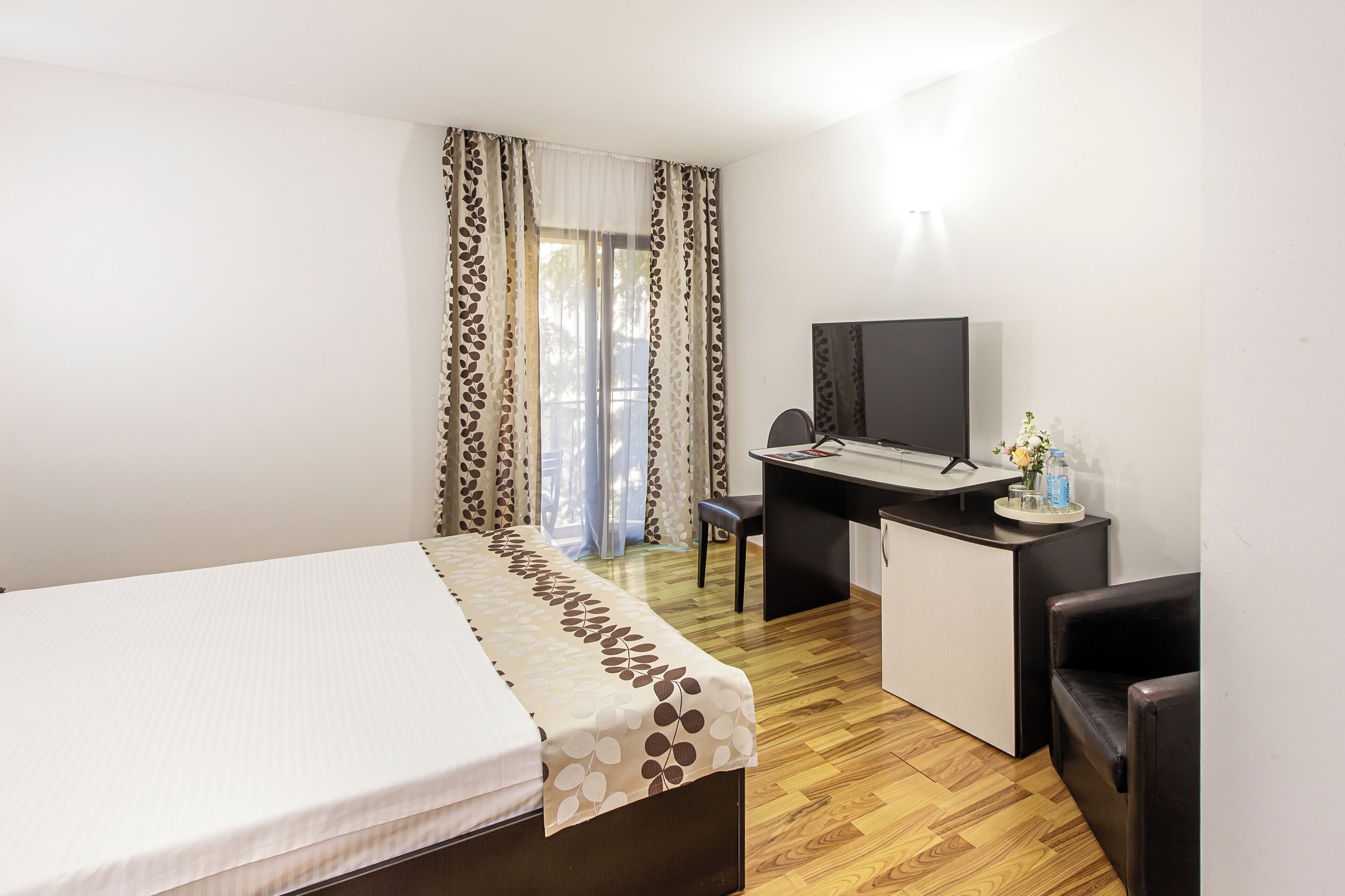 A hotel room with a bed, desk, chair, TV, and a small table with a vase and water bottles.