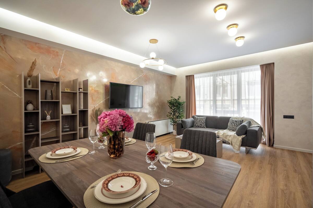Una sala de estar moderna con comedor, piso de madera, televisor de pantalla plana y un ventanal con cortinas.