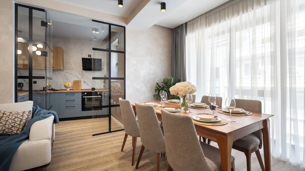 A modern dining area with a wooden table set for six, surrounded by chairs. A kitchen is visible through a glass partition to the left. - Manu Art Living (Bucharest)