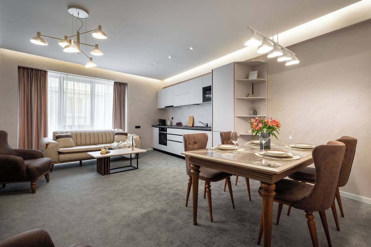 Una sala de estar moderna con sofá, mesa de comedor y sillas, una cocina al fondo y un ventanal con cortinas.