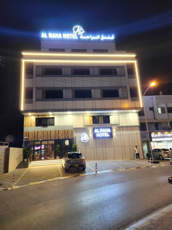 Exterior del Hotel Al Raha con un diseño moderno, destacando un llamativo letrero iluminado y un coche estacionado al frente.