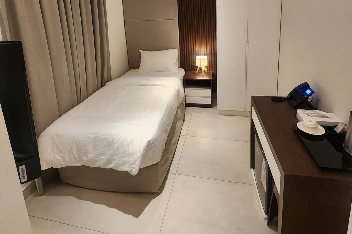 A hotel room with a bed, a desk with a phone, an air conditioner, a nightstand with a lamp, and a wardrobe.