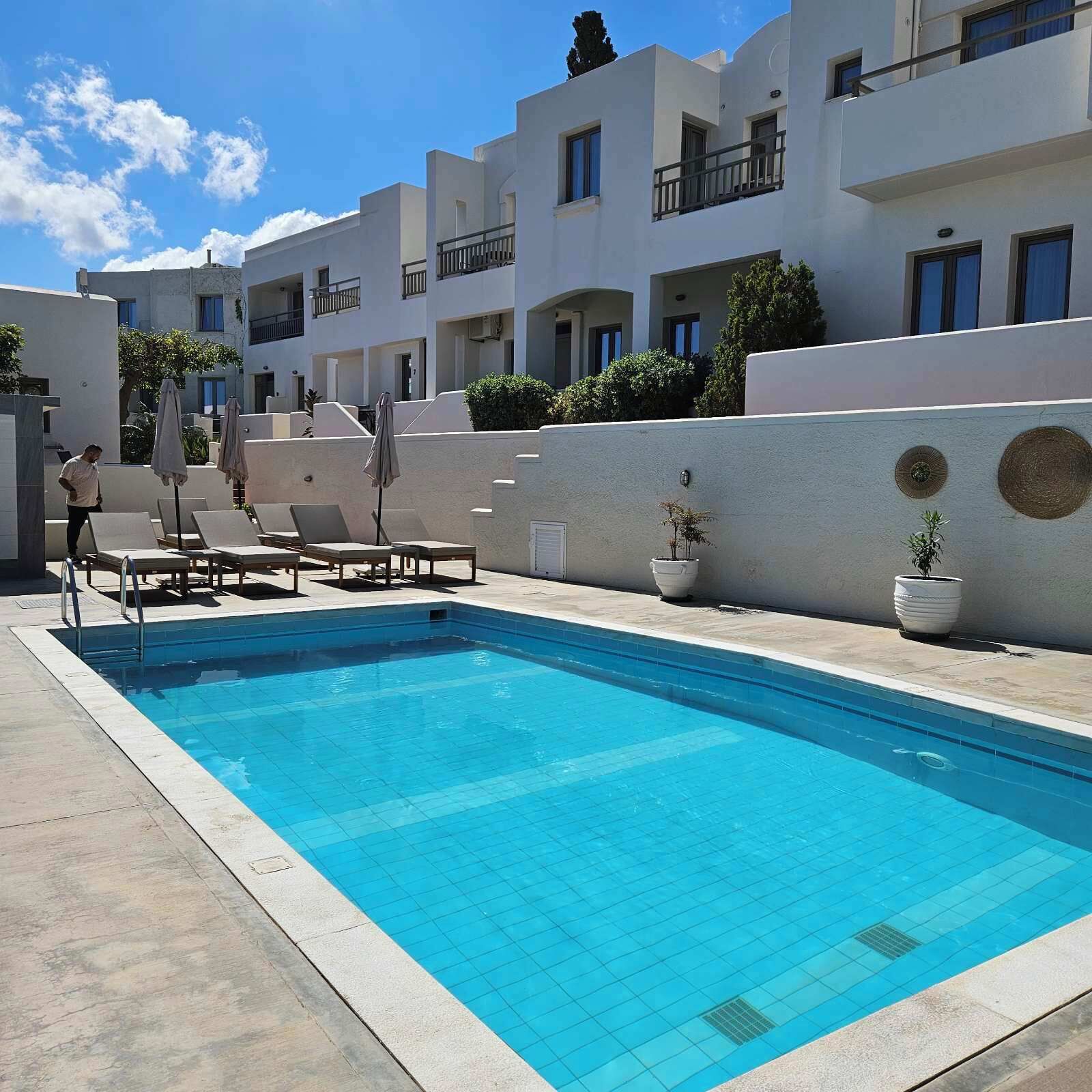 Une piscine entourée de bâtiments blancs dotés de balcons, avec des chaises longues et des parasols à proximité.