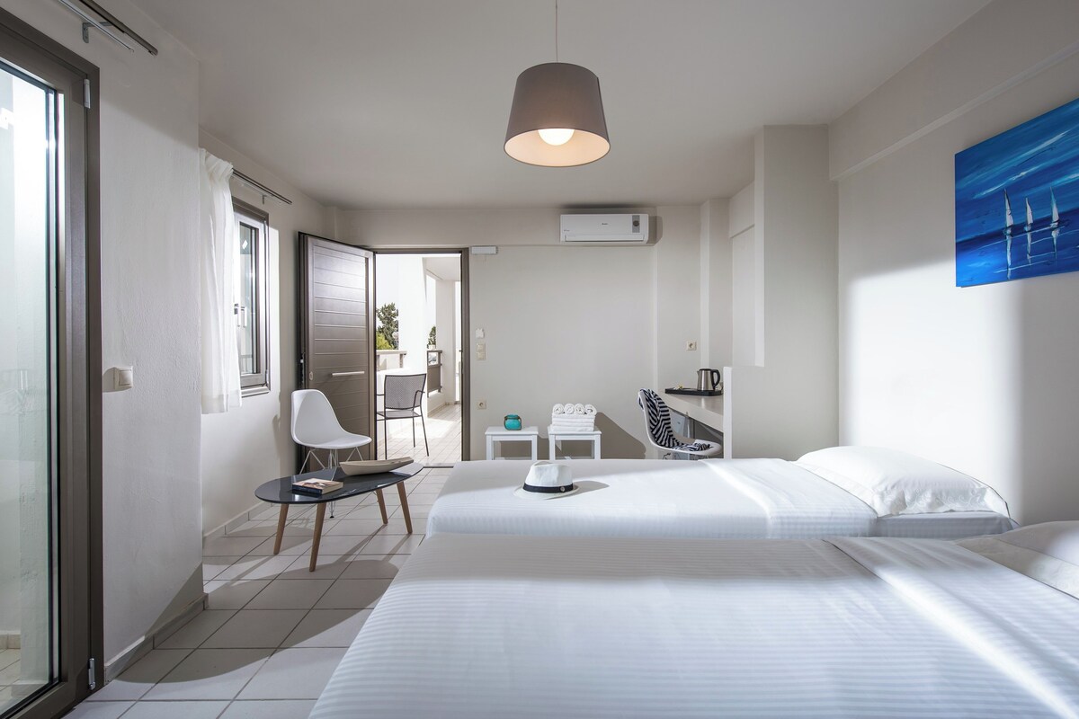 Une chambre d’hôtel avec deux lits, un coin salon avec une table et des chaises, un tableau au mur et une porte coulissante donnant sur un espace extérieur.