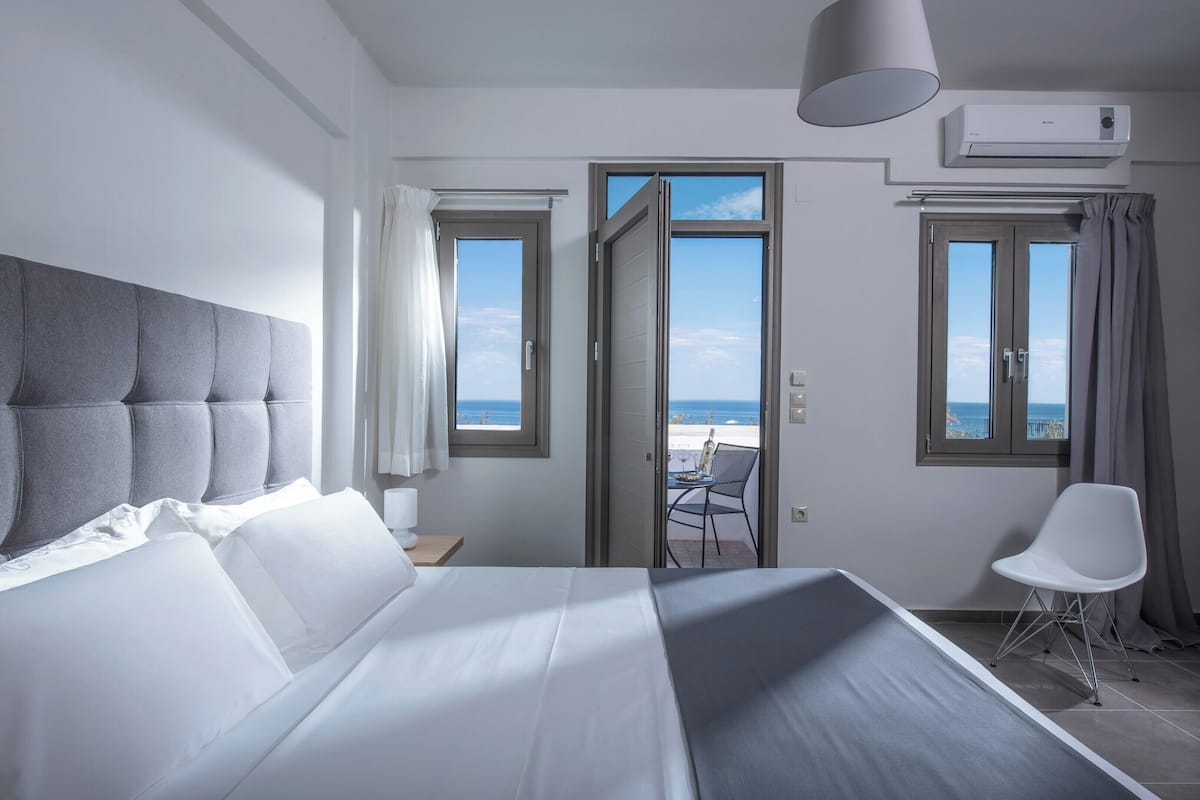 A hotel room with a large bed, a view of the sea through open doors, a balcony with a table and chair, and an air conditioning unit on the wall.