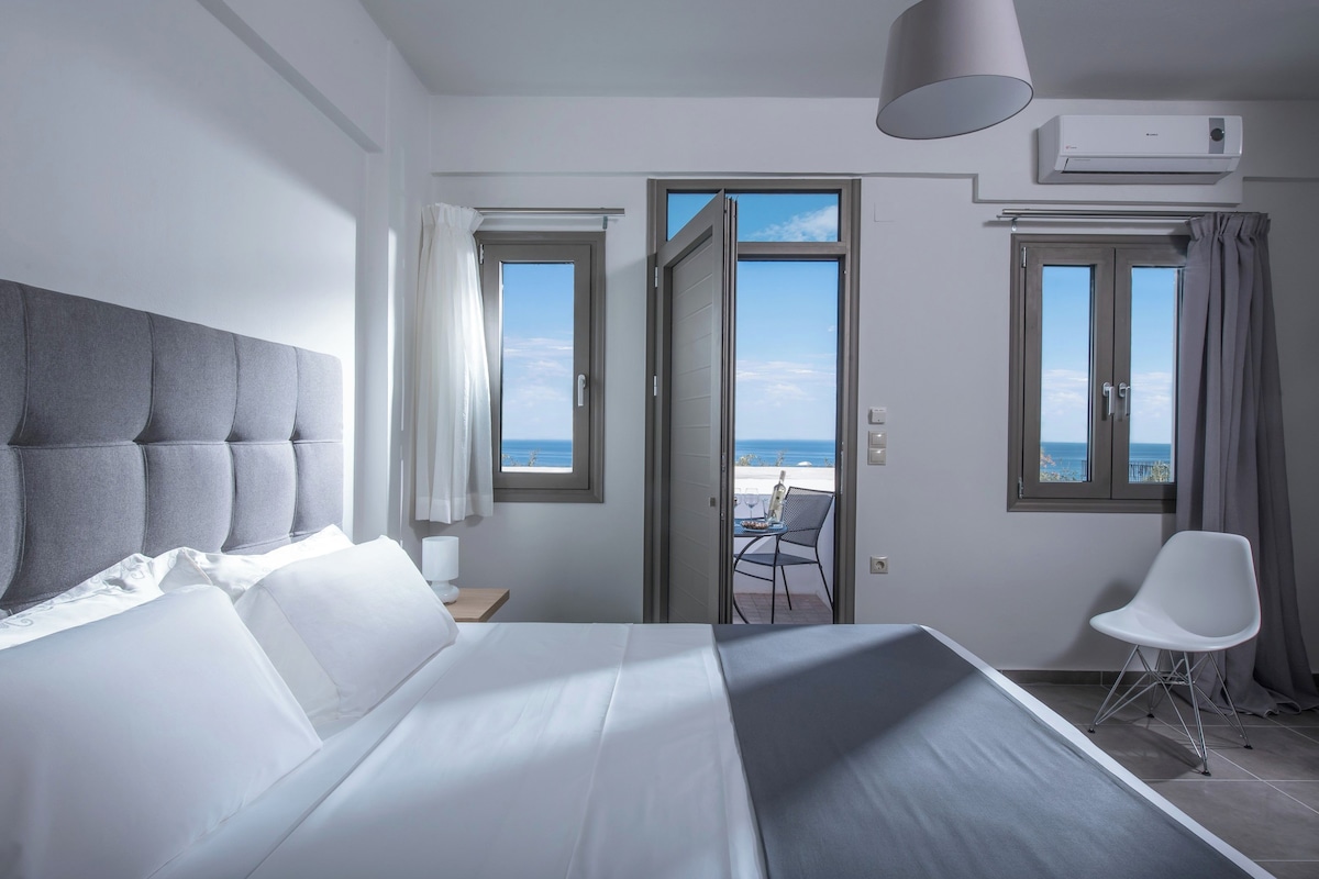 A hotel room with a large bed, a view of the sea through open doors, a balcony with a table and chair, and an air conditioning unit on the wall.