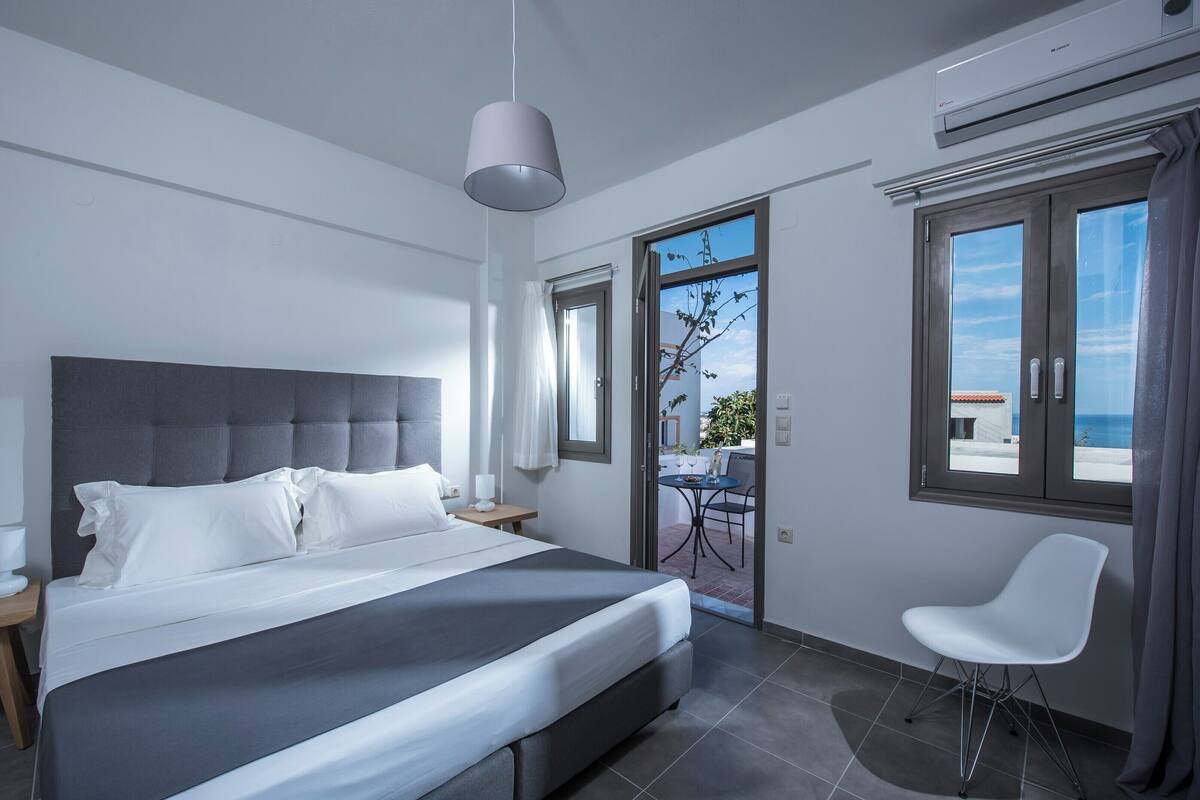 A modern hotel room with a large bed, a bedside table, a chair, and a view of the outdoors through a glass door.