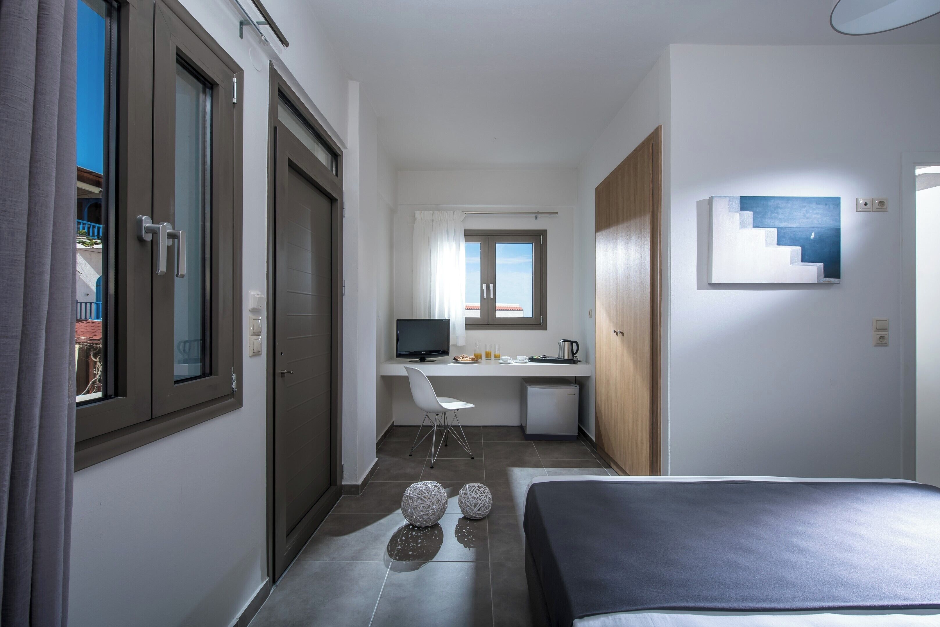 A compact hotel room with a bed, a desk with a computer, a chair, and a small kitchenette.