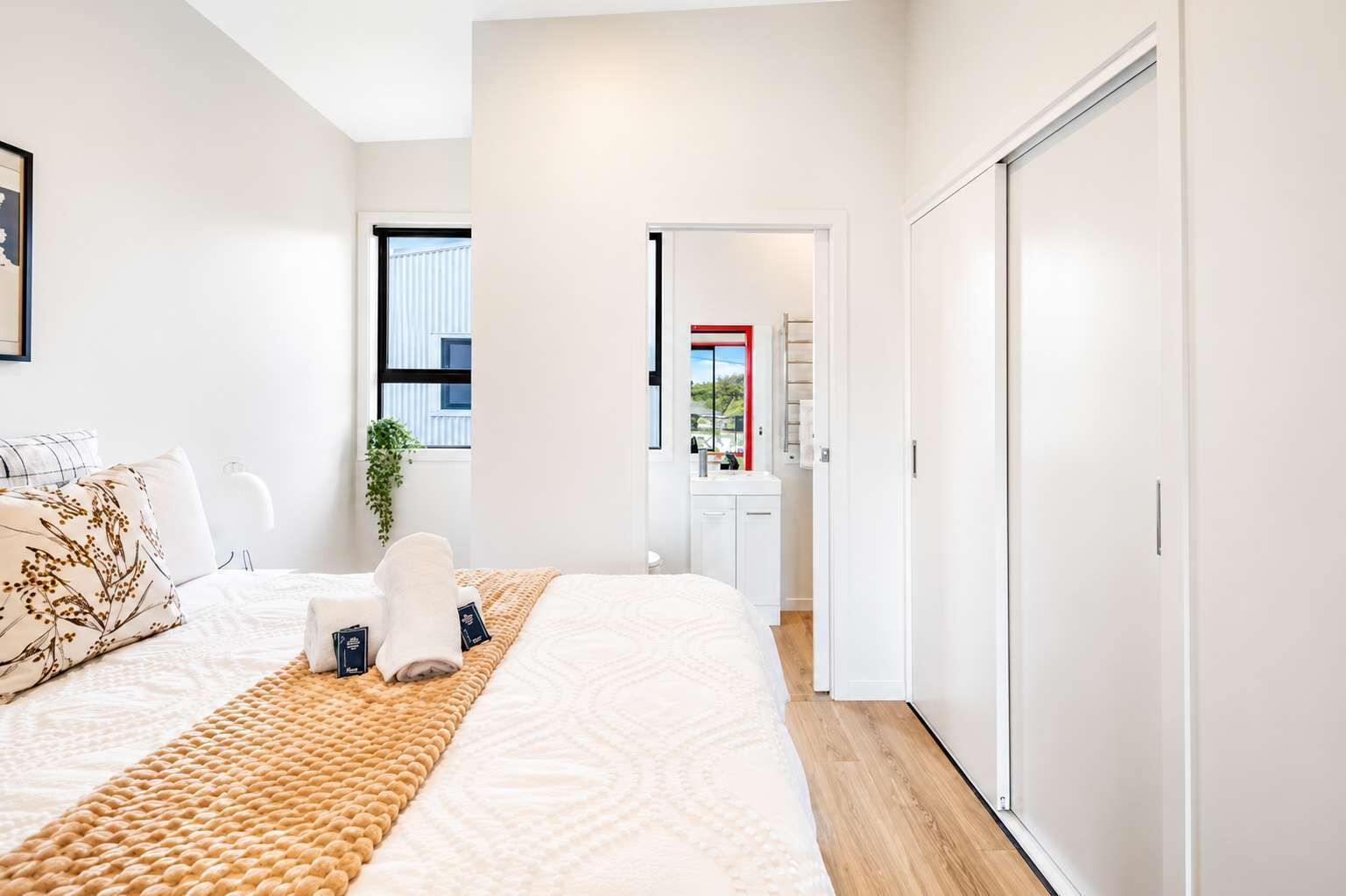 A modern bedroom with a large bed, a bedside table, a window with blinds, and a view of the outside.