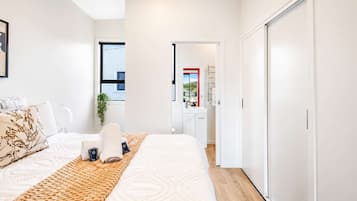 A modern bedroom with a large bed, a bedside table, a window with blinds, and a view of the outside.