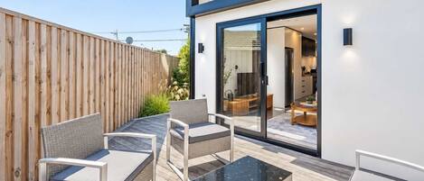 A modern outdoor patio with wicker furniture, a glass table, and a view into the house.