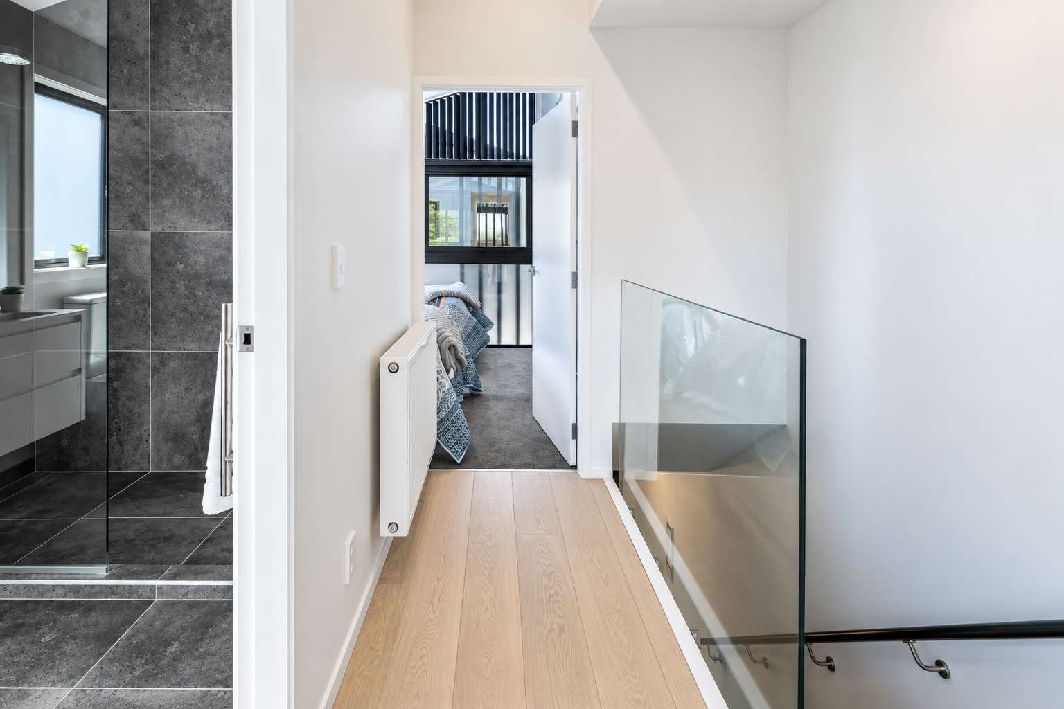 A modern interior with a hallway leading to a bedroom, a bathroom with a shower, and a glass railing.