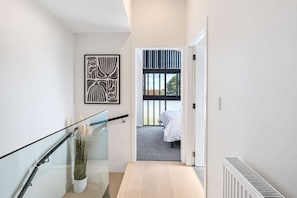A modern hallway with a glass railing, a potted plant, and a bed visible through a window.