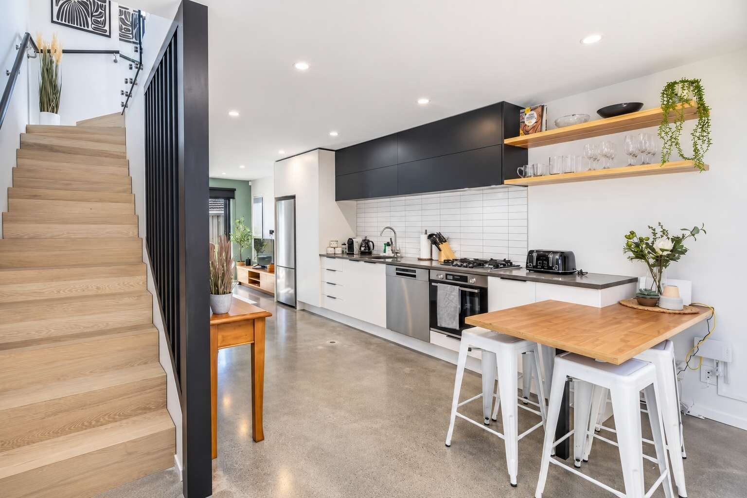 A modern kitchen with a wooden staircase, white cabinets, and a dining area with stools.