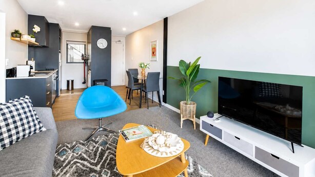 A modern living room with a sofa, a blue chair, a wooden coffee table, a TV, and a dining area with chairs and a table. - Newtown Tranquility | 2BR Wellington | Kitchen (Wellington)