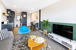 A modern living room with a sofa, a blue chair, a wooden coffee table, a TV, and a dining area with chairs and a table.