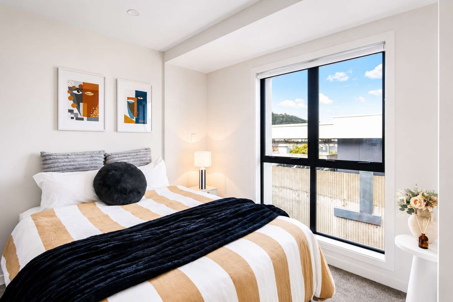 A hotel room with a large window, a bed with striped bedding, a black throw, and two abstract paintings on the wall.