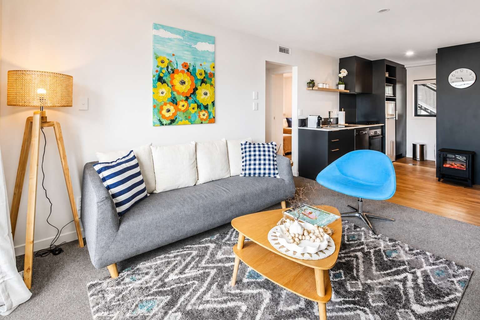 A modern living room with a sofa, a blue chair, a coffee table with a magazine, a painting, and a kitchen area in the background.