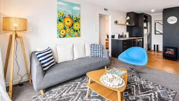 A modern living room with a sofa, a blue chair, a coffee table with a magazine, a painting, and a kitchen area in the background.