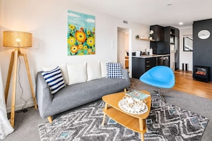 A modern living room with a sofa, a blue chair, a coffee table with a magazine, a painting, and a kitchen area in the background.