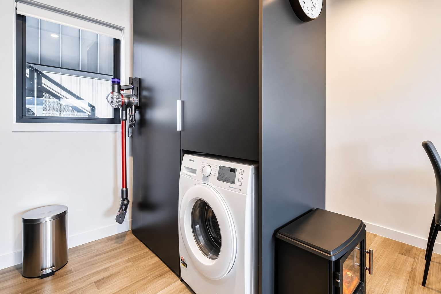 A compact laundry room with a front-loading washing machine, a small trash bin, and a heater.
