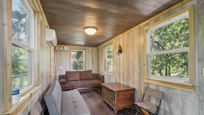 A cabin with a wooden interior, a brown sofa, a wooden chest, and a window overlooking greenery. - Fishing Pond & Treehouse: Tranquil Maysville Cabin (Maysville)