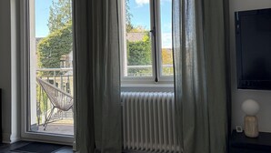 Une pièce avec une grande fenêtre, un radiateur, un téléviseur posé sur un meuble et une chaise placée à l’extérieur, devant la fenêtre.