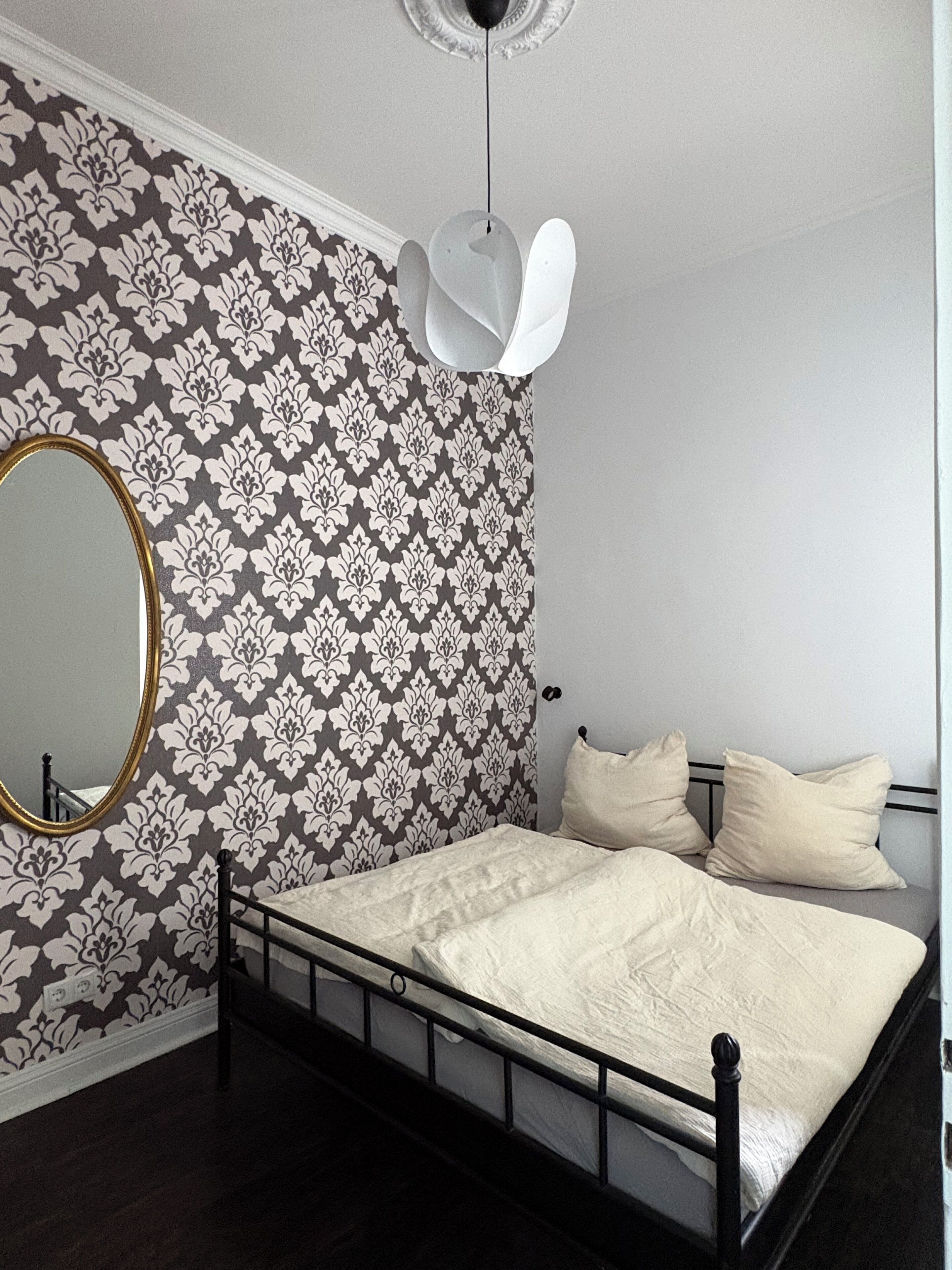 Une chambre avec un papier peint à motifs, un miroir rond, un lit avec une literie blanche et un luminaire suspendu.