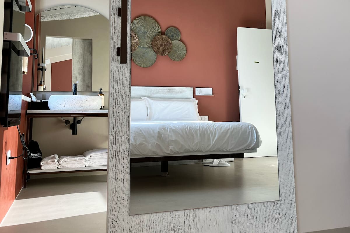 A modern bedroom with a large mirror, a bed, a white dresser, and a bathroom with a sink and mirror.