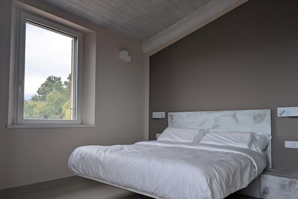 A bedroom with a large window, a bed with white bedding, and a wooden ceiling.