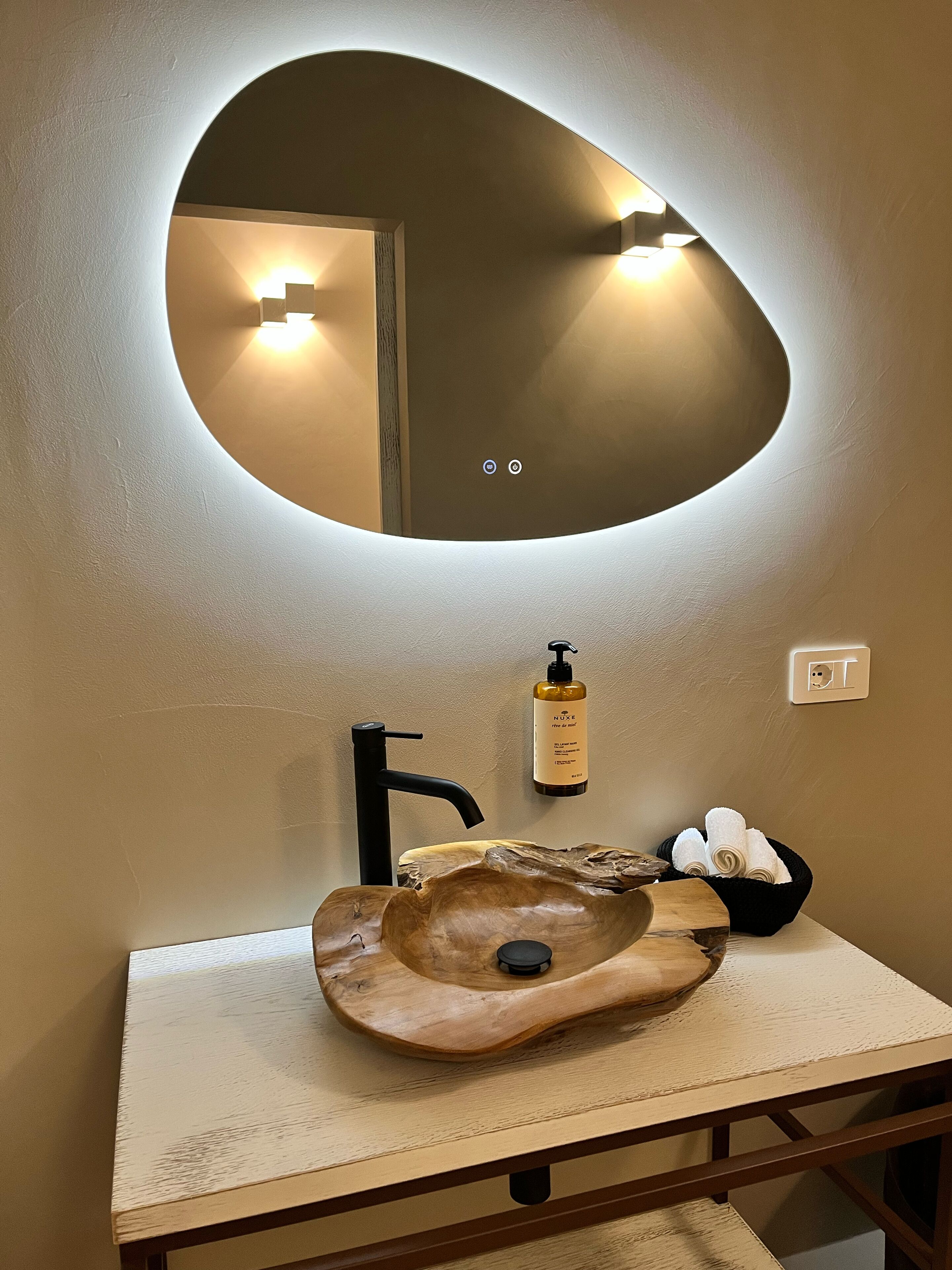 Une salle de bain avec un lavabo en bois, un robinet noir, un miroir rond avec éclairage intégré, une bouteille de gel douche Nuxe et un porte-serviettes.