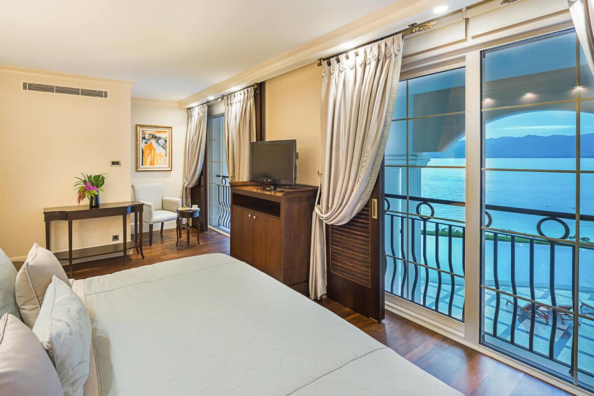 Habitación de hotel con una cama grande, un escritorio con televisor, una silla, una mesa con planta y un balcón con vista al mar.